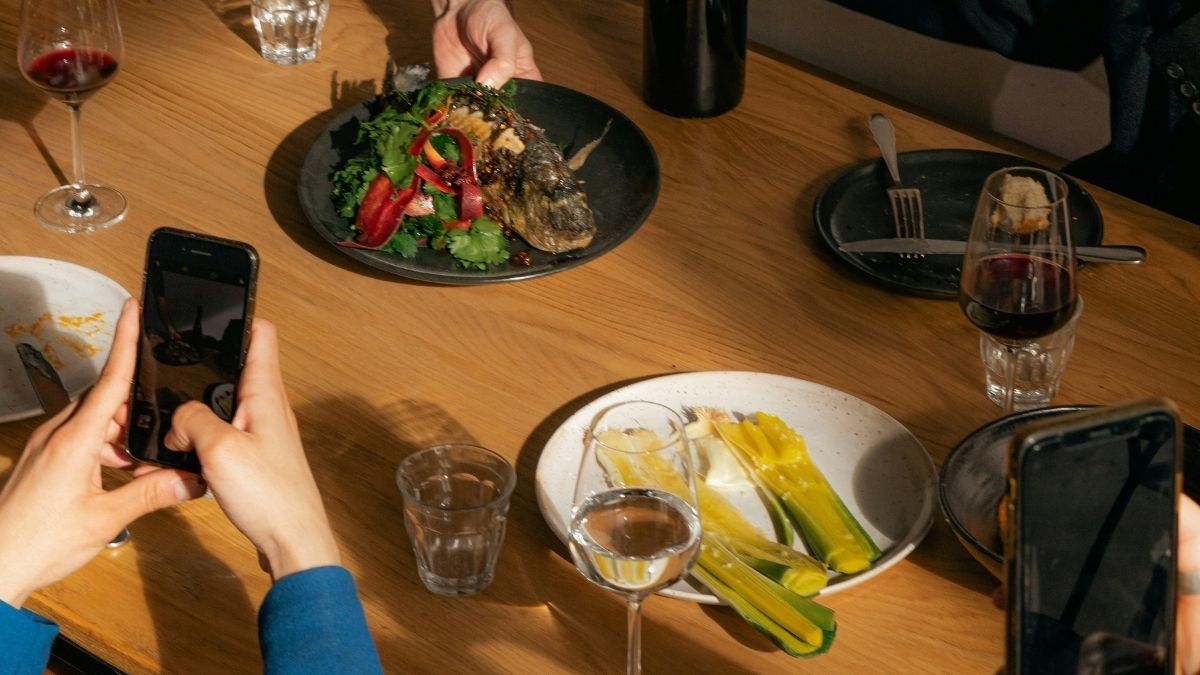 Person photographing restaurant dishes with a smartphone during a group meal, with wine glasses and plates visible from above